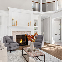 Traditional style living room using different types of moulding with 2 grey single cushion sofas on each side of a lit fireplace, a cream 4 seater sofa to the left, and a grey rectangle rug under a wood grain metal rectangle coffee table with metal vase on top that holds orange and yellow leaf branches in the center