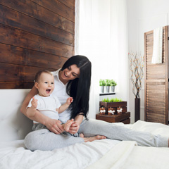 Woman holding her smiling baby while sitting on a bed with a white comforter and headboard and Pakari shiplap on the wall behind them