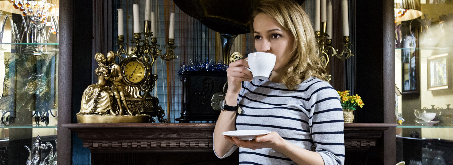 A woman with blonde hair wearing a grey and black striped sweater is drinking tea out of a white tea cup in her right hand and holding the tea plate in her left. She is in front of an elegant dentil mantel fireplace