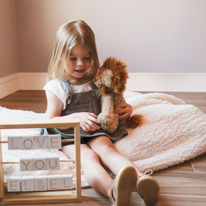 A little girl is sitting in a room with baseboard moulding on a blanket on the floor while holding a stuffed lion. There is a sign in front of her made out of blocks that reads 'Love you El & El'.