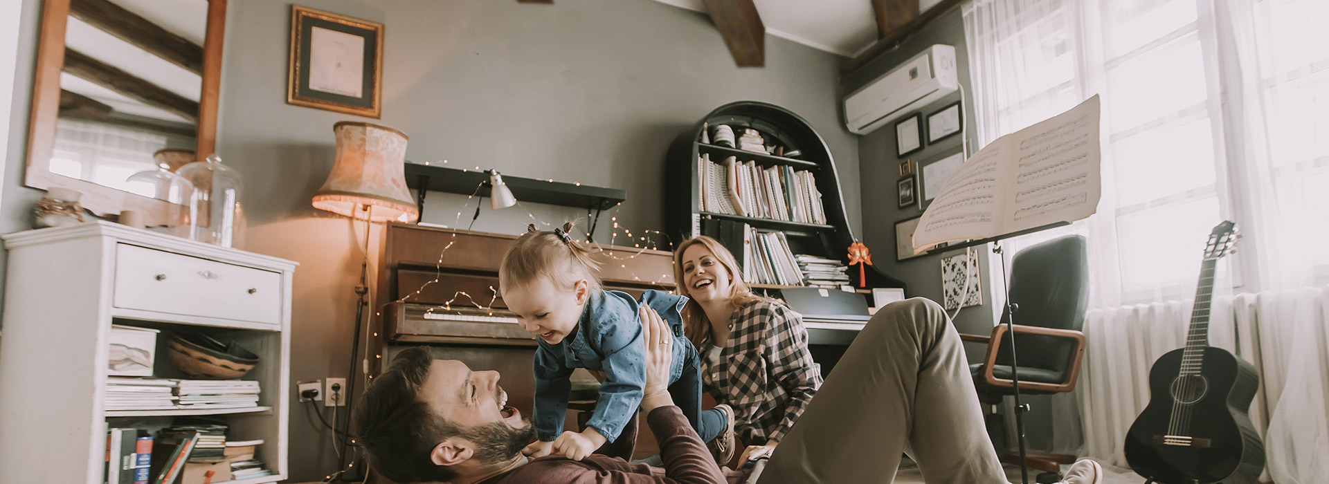 Family music room with a father laying on his back while he holds his young daughter horizontally and the mother is also on the floor sitting behind them. They are all smiling and laughing.