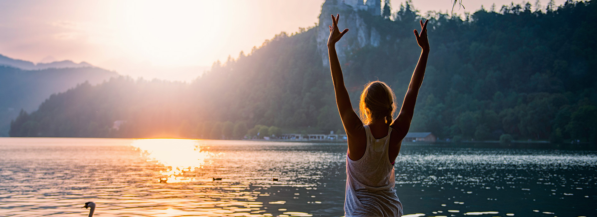 A photo taken behind a woman with both of her arms up towards the sky into a yoga pose in front of lake during the sunset