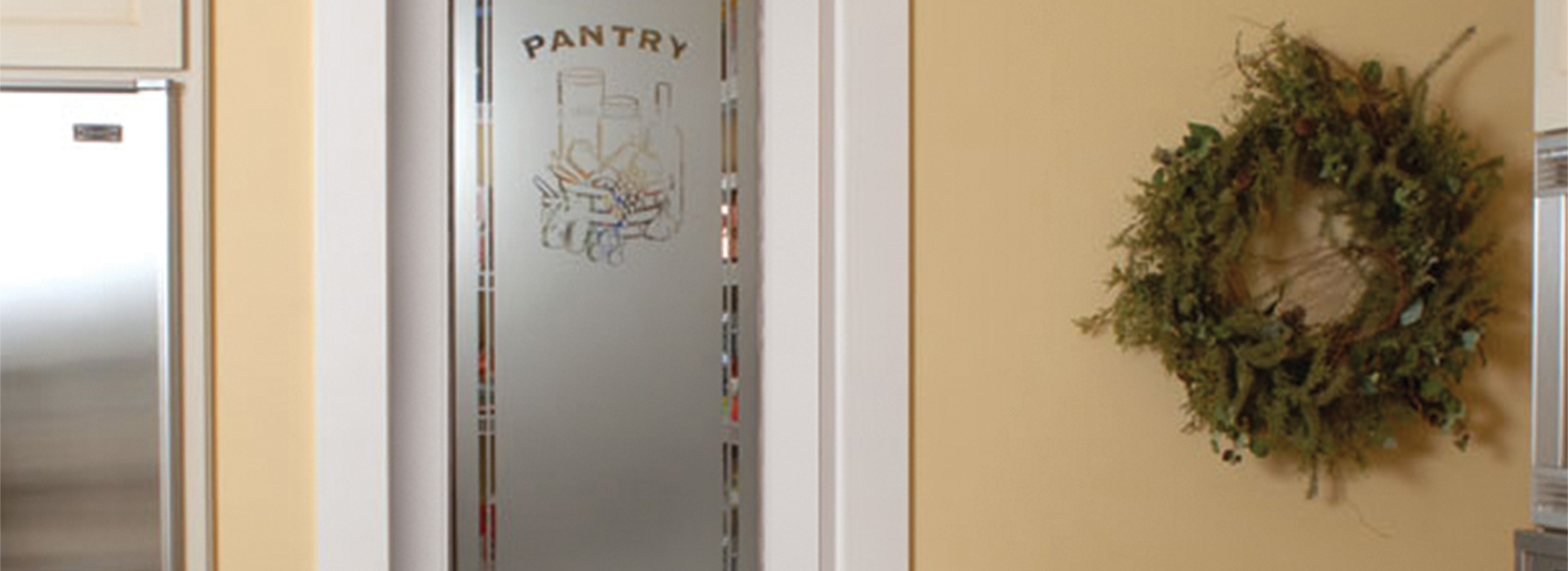 Close up shot of a yellow painted kitchen with an MDF primed decorative glass pantry door in the center, stainless steel fridge to the left, and green wreath to the right