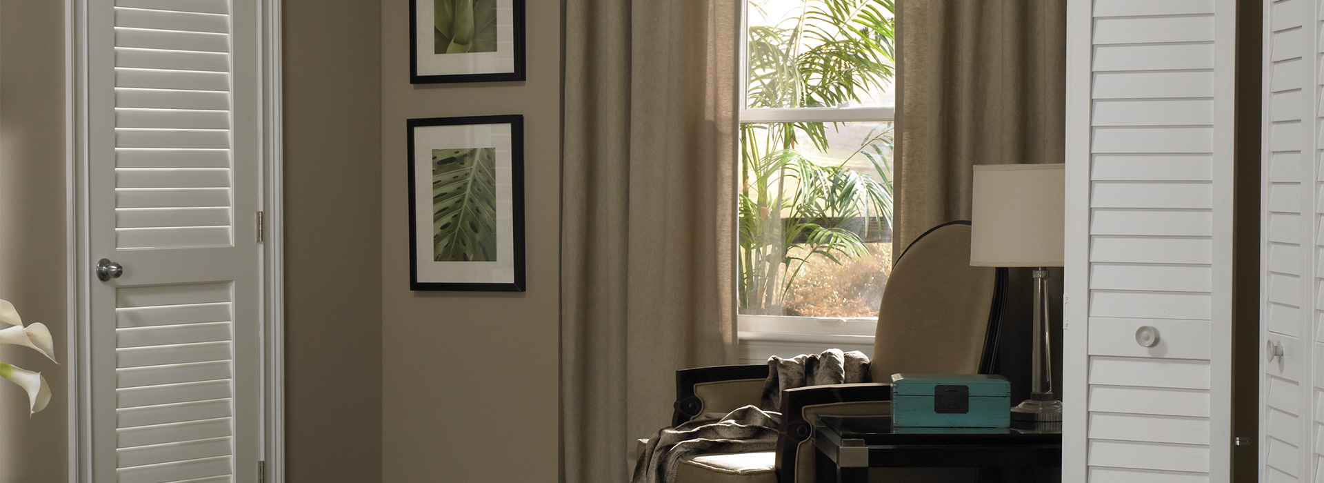 Coastal style living area with a MDF primed plantation shutter Louver door to the left, 2 photos of tropical leaves on the wall, and a single cushion chair to the right