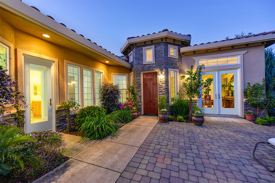 Mediterranean style home with a stone pathway and lots of greenery, 1 full lite white exterior door on the left side for a restroom, an 8 panel wood grained exterior door for the main entrance in the middle, and full lite white double doors tp the right leading to the dining room