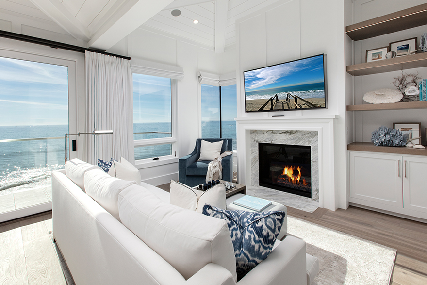 Coastal style living room with a beach window view to the left, white couch and ottoman facing a mounted flat screen tv and fireplace, a blue single cushion sofa in the left corner, and shelves to the far right 
