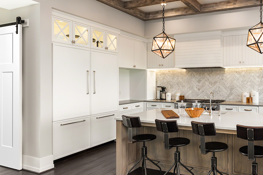 Craftsman style kitchen with white drawers and cabinets, white center island countertop with 4 dark metal stools, grey countertops flush with the wall and appliances, 2 geometric lights over the island, polyurethane beams on the ceiling, and a barn door on black tracks