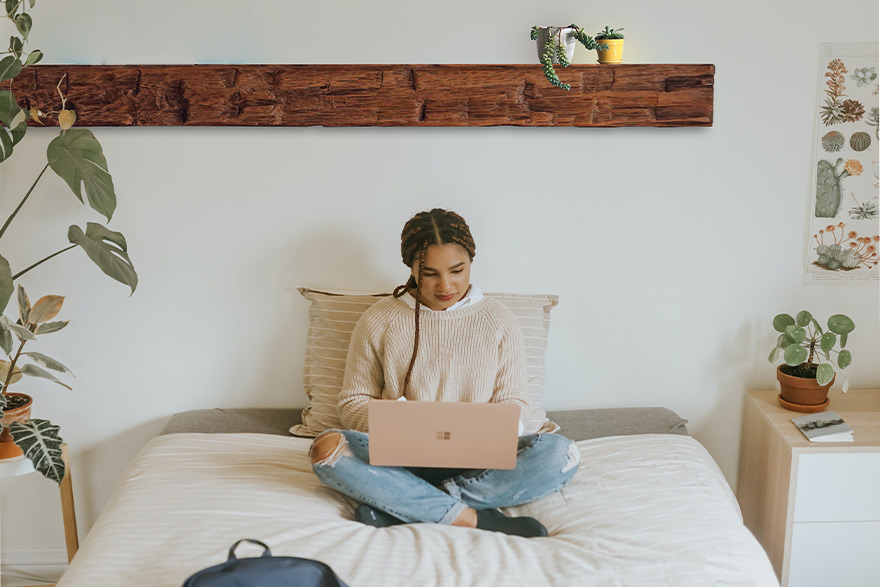 Woman sitting on a bed while typing on a laptop that is on her lap and a beam used as a shelf to hold 2 potted plants is over her head 