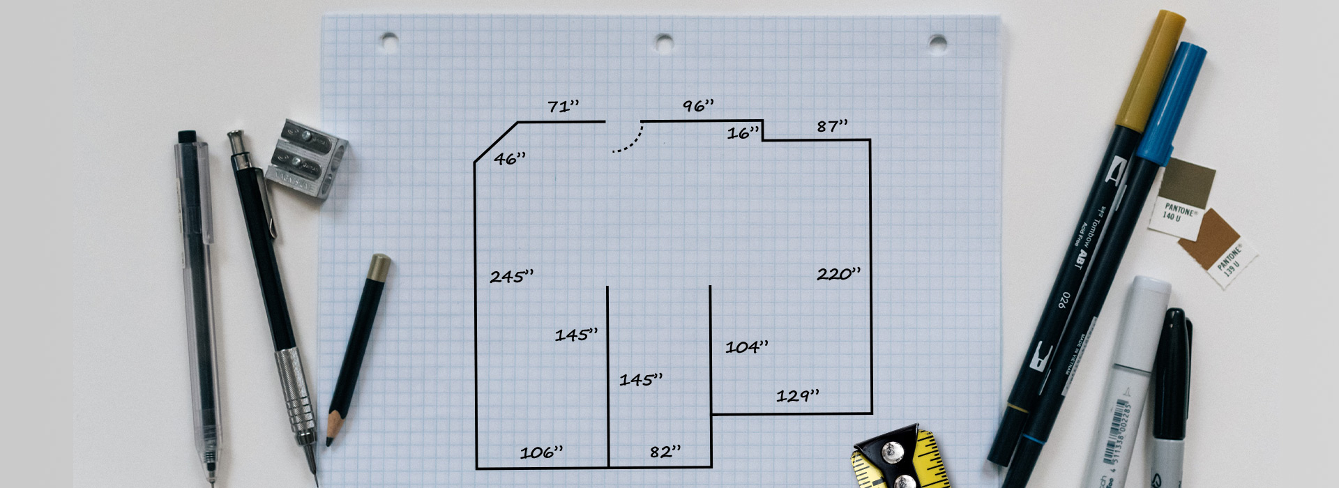 Graph paper with an outline of a house floor plan with different measurements for each side and pencils, pens, and markers scattered around it