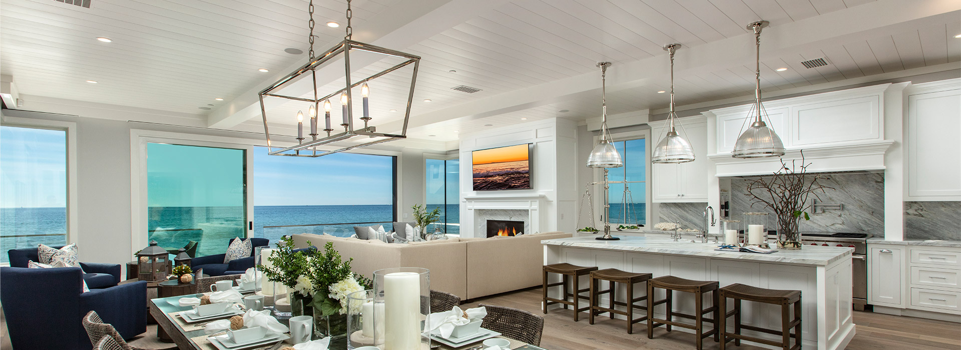 Coastal style living with a white shiplap ceiling that has sliding glass doors on the left that lead to the patio with an ocean view, in the bottom left corner of the room there are 3 blue single cushion sofas, in the top left corner there is an L shaped cream couch, 2 grey single cushion sofas, a fireplace, and a mounted flat screen tv, on the bottom right corner there is a set 8 seater rectangle wooden dining table and to the top right is a white marble kitchen with an island with 4 brown stool and 3 glass lights overhead.